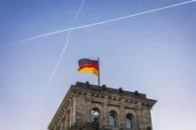 Ein Gebäudeturm des Reichstags in Berlin mit wehender deutscher Flagge. Am Himmel kreuzen sich zwei Kondensstreifen.